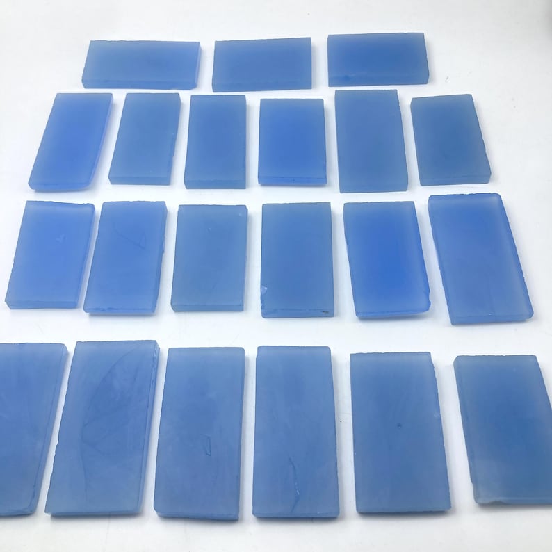 May include: A collection of light blue rectangular glass tiles are arranged in rows. Each tile has a slightly irregular shape and a smooth, translucent appearance. The color is a soft, muted blue. The tiles are displayed from an overhead perspective.