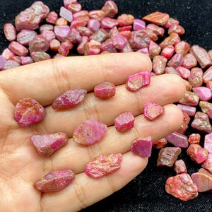 May include: A collection of raw, uncut ruby gemstones in varying sizes and shapes. The gemstones display a range of pink and red hues, with some showing a translucent quality. The rough texture and natural forms of the stones are visible.