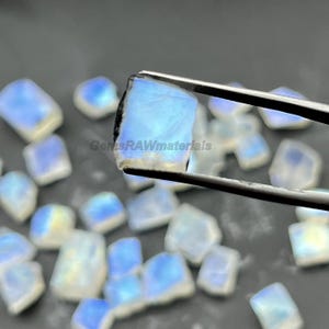 May include: A collection of raw, translucent moonstone gemstones. One stone is held by tweezers, showcasing its blue and white iridescence. The background features additional moonstones, creating a display of natural mineral specimens. The text "GemsRAWmaterials" is visible.