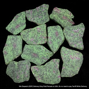 Könnte beinhalten: Eine Ansammlung von zehn unregelmäßigen, flachen Stücken aus Zoisit-Mineral mit einer gesprenkelten grün-schwarzen Basis und eingebetteten Flecken aus tief magentafarbenem Rubin. Die Steine haben raue, gezackte Kanten und eine natürliche Oberfläche.