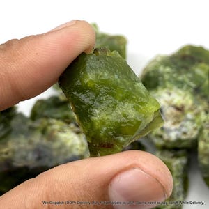 May include: A raw, translucent olive green mineral specimen with an irregular, jagged shape and a glossy, crystalline texture. The stone displays deep green hues with subtle variations in clarity and small dark inclusions throughout its surface.