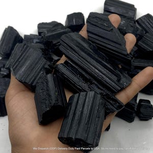 Peut inclure: Une collection de cristaux de tourmaline noire brute présentant des stries verticales distinctes et des bords irréguliers. Ces spécimens minéraux opaques et sombres sont regroupés, mettant en valeur leur structure cristalline naturelle, non polie et colonnaire.