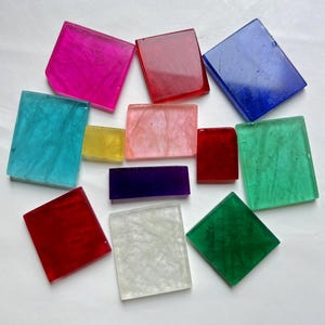 Puede incluir: Un surtido de baldosas de vidrio coloridas, translúcidas, cuadradas y rectangulares en tonos de rosa, rojo, azul, turquesa, amarillo, melocotón, morado y verde. Las baldosas están dispuestas sobre una superficie blanca.