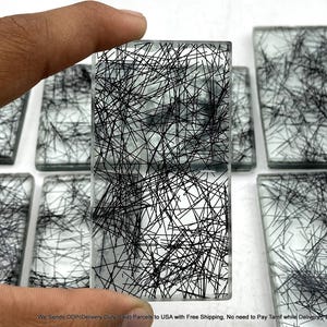 Pode incluir: Um cabochão retangular de vidro transparente com um padrão interno de finas inclusões pretas em forma de agulha que se cruzam. A peça tem uma superfície lisa e plana com bordas nítidas, exibindo uma disposição densa e caótica de linhas escuras.