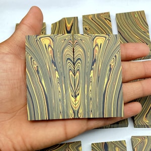 May include: Two rectangular pieces of material with a swirling pattern of yellow, brown, and black. The pieces are held in a hand, with other similar pieces visible in the background. The pattern resembles wood grain.