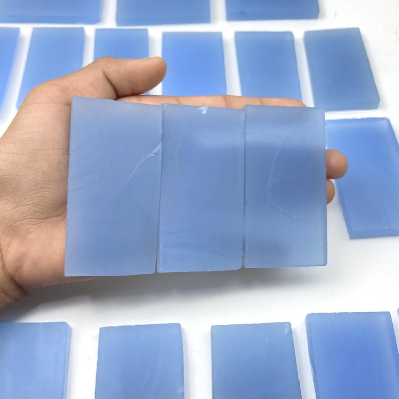 May include: A collection of rectangular, pale blue gemstone slabs. The slabs are held in a hand, with more visible in the background. The stones have a smooth, translucent appearance, and are likely used for jewellery or crafting.