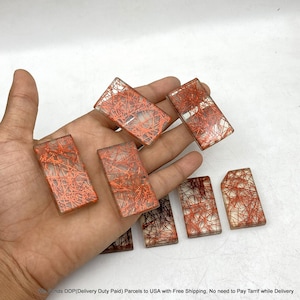 May include: A collection of seven rectangular clear resin cabochons featuring embedded orange and red needle-like inclusions. Each piece has a smooth, polished surface and a distinct pattern of crisscrossing metallic-coloured fibres within the material.
