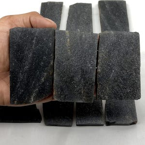 Puede incluir: Una colección de baldosas de piedra rectangulares de color gris oscuro con una superficie texturizada y moteada. Las piedras tienen bordes rugosos e irregulares y un acabado mate, dispuestas en cuadrícula con algunas piezas sostenidas en la mano.