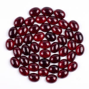 Può includere: Una collezione di numerosi cabochon sciolti di forma ovale in un colore rosso granato profondo e traslucido. Ogni pezzo presenta una superficie liscia, lucida e arrotondata, disposta in un denso gruppo circolare su sfondo bianco.