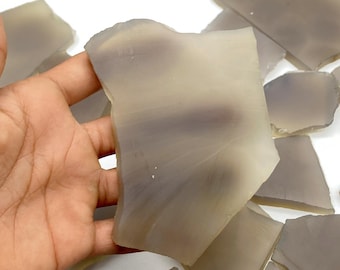 May include: A collection of translucent, irregularly shaped, pale gray gemstone slices. The stones have a smooth, polished appearance with subtle color variations. The image shows the stones in a hand, with others scattered in the background.
