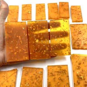 May include: Several rectangular, amber-colored resin blocks with gold leaf flakes are displayed. The blocks vary in size and are arranged on a white surface. The resin is translucent, allowing light to pass through, and the gold flakes add a decorative element.