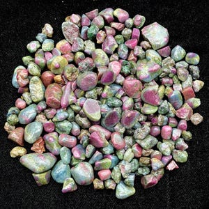 Op de afbeelding: Een verzameling getrommelde edelstenen in verschillende maten en vormen. De stenen vertonen een mix van kleuren, waaronder roze, rood, groen en blauw. De edelstenen zijn verspreid op een zwart oppervlak.