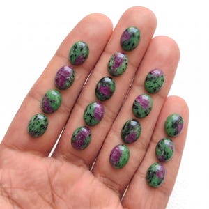 May include: A collection of fifteen oval-shaped ruby zoisite cabochons, each featuring a distinct mottled pattern of vibrant magenta-pink ruby inclusions set against a deep forest green zoisite background with dark black mineral speckles.