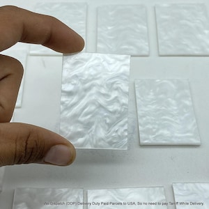 Puede incluir: Una baldosa rectangular blanca con un patrón nacarado arremolinado se sostiene entre los dedos. Varias baldosas similares están dispuestas en el fondo. Las baldosas tienen un acabado brillante y un diseño ondulado.