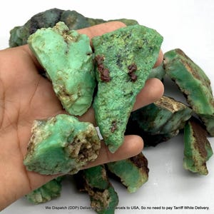 May include: A collection of raw, unpolished chrysoprase stones featuring a vibrant mint green colour with natural brown and dark inclusions. These rough mineral specimens have irregular, jagged edges and a matte, textured surface appearance.