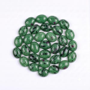 May include: A collection of oval-shaped cabochons made of green zoisite with black hornblende inclusions and small, distinct magenta-coloured ruby spots. Each polished gemstone features a smooth, rounded surface and a glossy, reflective finish.