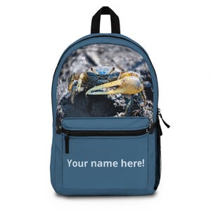 Op de afbeelding: Een blauwe rugzak met een fotografische print van een krab op het bovenste gedeelte en de tekst "Your name here!" in het wit op het voorvak. De tas heeft een zwart handvat aan de bovenkant, zwarte ritsen en een zijvak van gaas voor opslag.