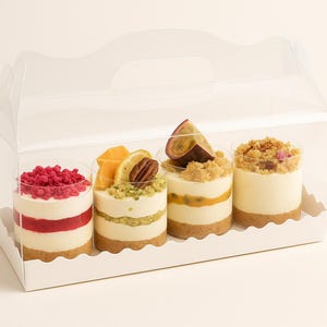 May include: Four individual desserts in clear plastic cups, each with a different topping. The desserts are arranged in a white, handled carrier. The toppings include raspberries, citrus fruits, a pecan, and passion fruit.