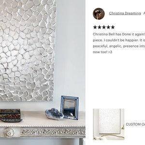 May include: A silver textured abstract painting with a raised, 3D effect. The painting is hanging on a white wall above a white console table with a decorative peacock figurine, a small picture frame, and a blue heart-shaped stone.