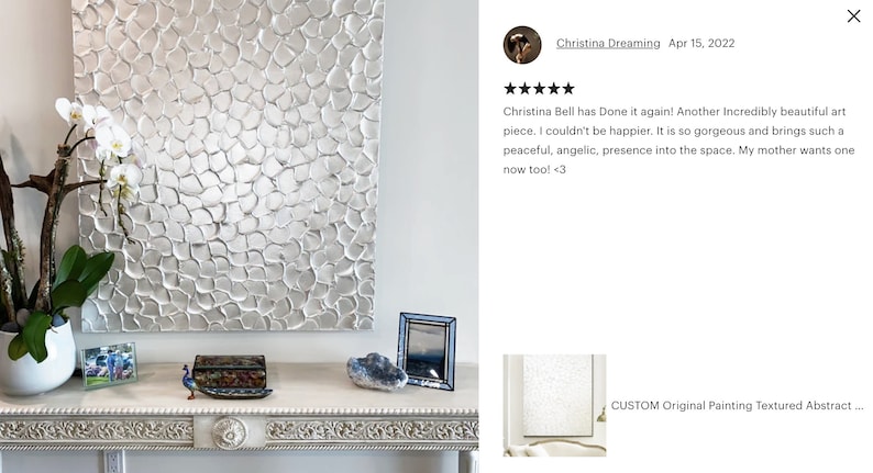 May include: A silver textured abstract painting with a raised, 3D effect. The painting is hanging on a white wall above a white console table with a decorative peacock figurine, a small picture frame, and a blue heart-shaped stone.