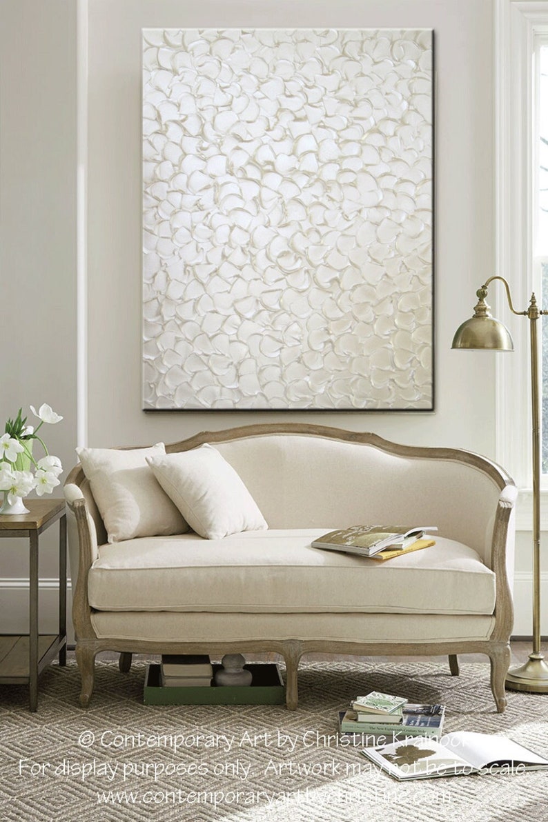 May include: A white abstract painting with a textured surface. The painting is hanging on a white wall above a white couch with pillows. The couch is in a room with a rug and a lamp.