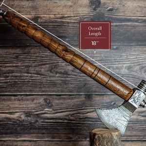 May include: A handcrafted tomahawk with a wooden handle and a metal head. The overall length is 45.7 cm. The handle is a dark brown color with a textured design. The metal head has an etched pattern and a silver finish.