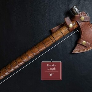 May include: A handcrafted axe with a wooden handle and a brown leather sheath. The handle is 40.6 cm long. The axe head is made of metal and has a sharp edge. The leather sheath is secured with brass-colored rivets.