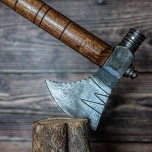 May include: A handmade axe featuring a wooden handle and a patterned steel blade. The axe head rests on a small wooden stump. The handle is dark brown, and the blade has a distinctive design.