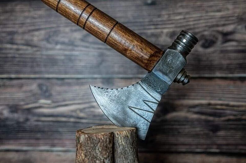 May include: A handmade axe featuring a wooden handle and a patterned steel blade. The axe head rests on a small wooden stump. The handle is dark brown, and the blade has a distinctive design.