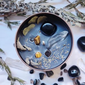 Op de afbeelding: Een ronde, donkerblauwe kaars in een koperkleurige blik, versierd met kristallen, stenen en gedroogde kruiden. De kaars heeft een heldere kwartskristal, tijgeroogstenen en zwarte stenen. Gedroogde bladeren en kruiden zijn verspreid.