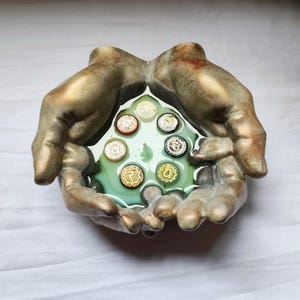 Op de afbeelding: Een bronzen sculptuur van twee handen die een kom vormen en een plasje lichtgroene vloeistof vasthouden. In de vloeistof bevinden zich zeven cirkelvormige, kleurrijke chakra-symbolen. De sculptuur heeft een verweerde, antieke uitstraling.