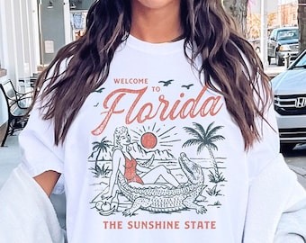 Könnte beinhalten: Weißes T-Shirt mit einem Grafikdruck einer Frau, die auf einem Alligator sitzt, mit dem Text "Willkommen in Florida, The Sunshine State".