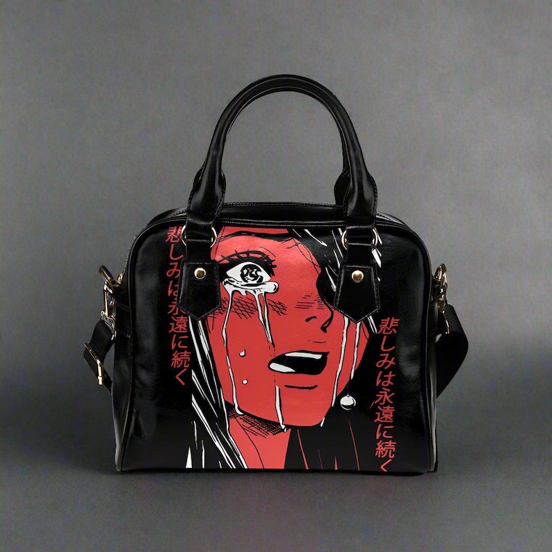 May include: Black handbag featuring a red and white manga-style illustration of a crying face. The bag has a top handle, a shoulder strap, and gold-colored hardware. Japanese text is printed on the side of the bag.