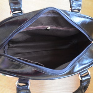 May include: Black handbag with a glossy finish, featuring a top zipper and two handles. The interior is lined with brown fabric. Silver-toned hardware accents the handles and zipper.