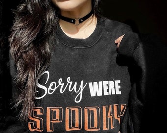 May include: A black sweatshirt with the text "Sorry Were Spooky" in white and orange lettering. The model is wearing a black choker necklace with a silver ring.