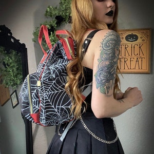 May include: Black backpack featuring a white spiderweb pattern, red detailing, and gold zips. The bag has black straps and a silver chain. A wooden sign with "Trick or Treat" is visible in the background.