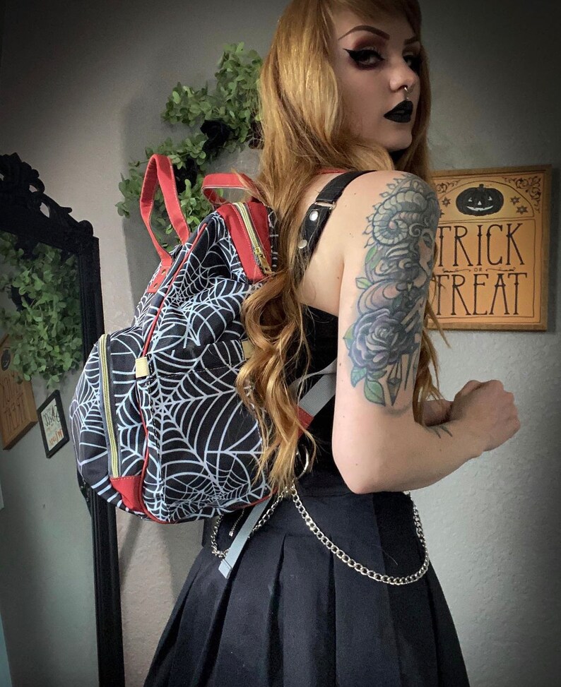 May include: Black backpack featuring a white spiderweb pattern, red detailing, and gold zips. The bag has black straps and a silver chain. A wooden sign with "Trick or Treat" is visible in the background.