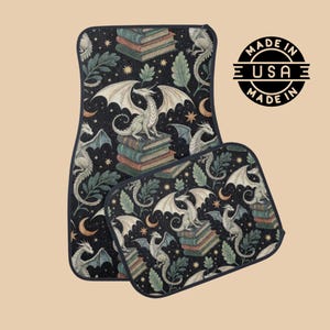 May include: Set of two car floor mats with a fantasy theme. The mats feature a black background with a repeating pattern of white dragons, stacks of books, leaves, stars, and moons. The mats are labeled "MADE IN USA".