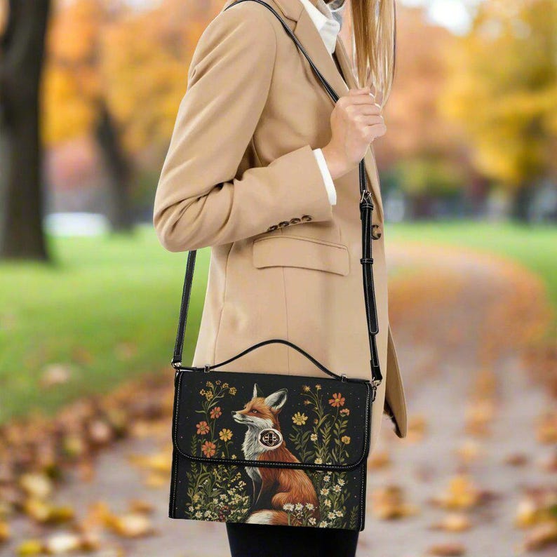 May include: A black crossbody bag with a fox illustration and floral design. The bag has a long adjustable strap and a top flap closure.