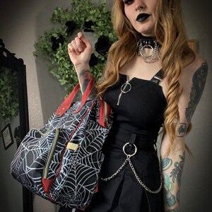 May include: Black and white spiderweb design backpack with red trim and gold zips. The bag has a top handle and a front pocket. The backpack is part of a gothic fashion ensemble.