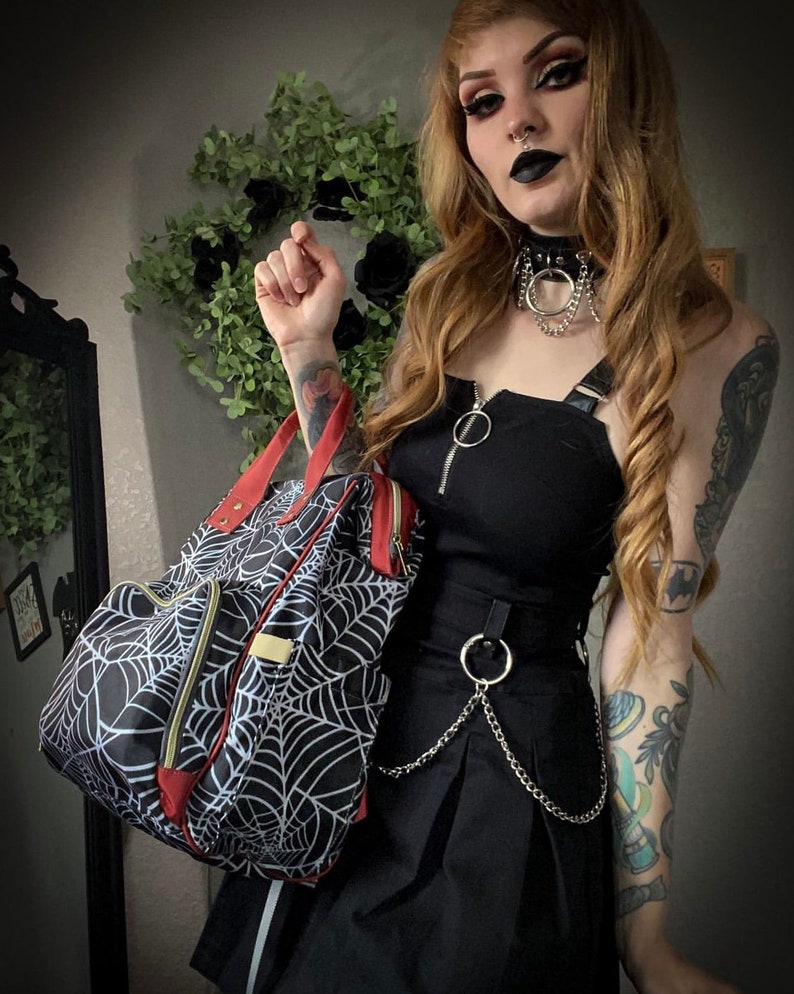 May include: Black and white spiderweb design backpack with red trim and gold zips. The bag has a top handle and a front pocket. The backpack is part of a gothic fashion ensemble.