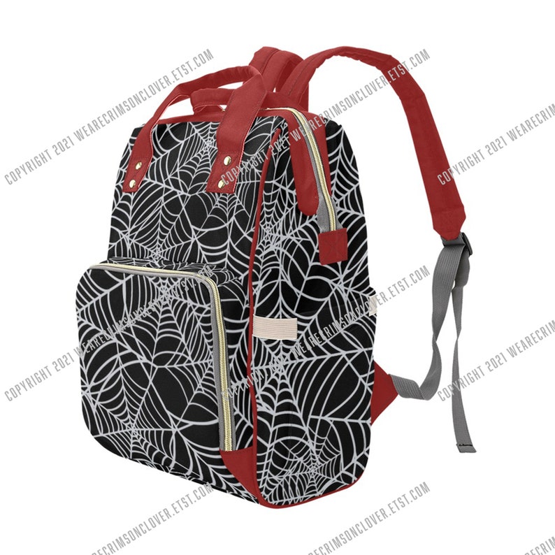May include: Black backpack featuring a white spiderweb design. The bag has red detailing on the top handle, straps, and base. Gold-coloured zips and grey straps complete the look. The text "COPYRIGHT 2021 WEARECRIMSONCLOVER.ETST.COM" is printed on the bag.