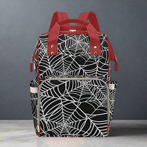 May include: Black backpack featuring a white spiderweb design. The bag has red straps and trim, including the top handle and shoulder straps. The bag has multiple pockets and a gold zip.