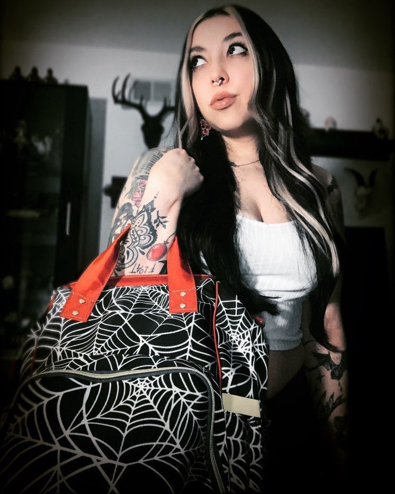 May include: A black and white tote bag featuring a spiderweb pattern, with orange handles. The bag has a zip closure. The bag is held by a person. A fashionable accessory.