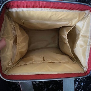 May include: Interior view of a red and grey tote bag, revealing multiple compartments and a light tan lining. The bag features a zipped pocket and grey handles at the top.