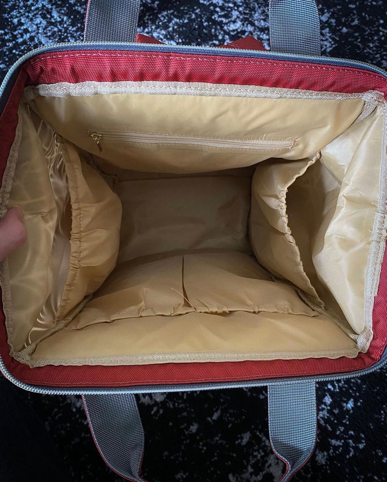 May include: Interior view of a red and grey tote bag, revealing multiple compartments and a light tan lining. The bag features a zipped pocket and grey handles at the top.