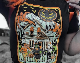 May include: Black t-shirt featuring a vintage-style illustration of a spooky house with a pumpkin-headed ghost and three trick-or-treaters. The illustration includes a devil, a witch, and a skeleton.