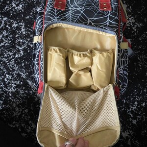 May include: Black and white spiderweb-patterned backpack with red detailing. The open bag reveals a tan interior with multiple pockets and a mesh compartment. The bag has a top handle and adjustable straps.