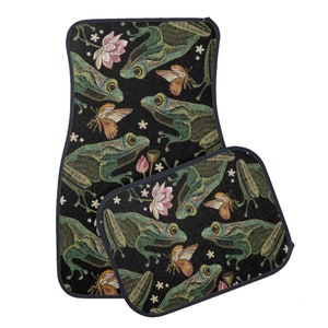Puede incluir: Un juego de dos alfombrillas para coche con fondo negro y un patr&oacute;n repetido de ranas verdes, flores rosas y mariposas. Las alfombrillas tienen un borde azul oscuro.
