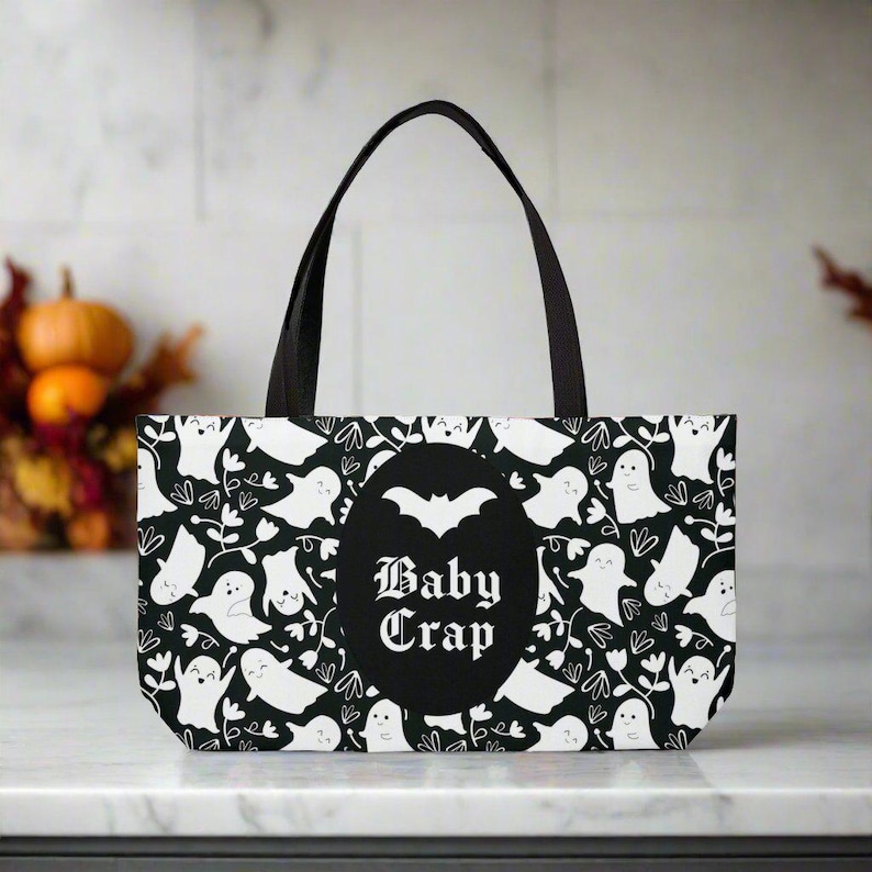May include: Black and white tote bag with a ghost and bat pattern. The bag has a black circle with the text "Baby Crap" in white.